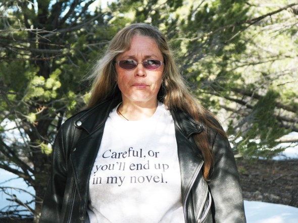Photo of upper body of author Kaye Lynne Booth in front of pine trees.Text on shirt: Careful, or you'll end up in my novel