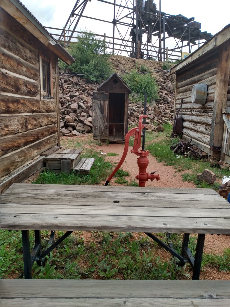 Wooden outhouse between two log cabins and a handpump and picnic table in foreground.