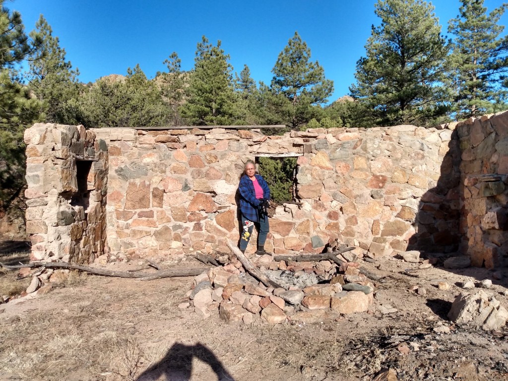 Kaye Lynne Booth in a blue overshirt in front of remains of old cabin, with trees and blue sky in the background
